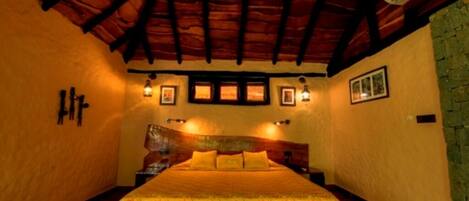 A cozy room with a wooden ceiling, a bed with a patterned bedspread, wall-mounted lights, and framed pictures.