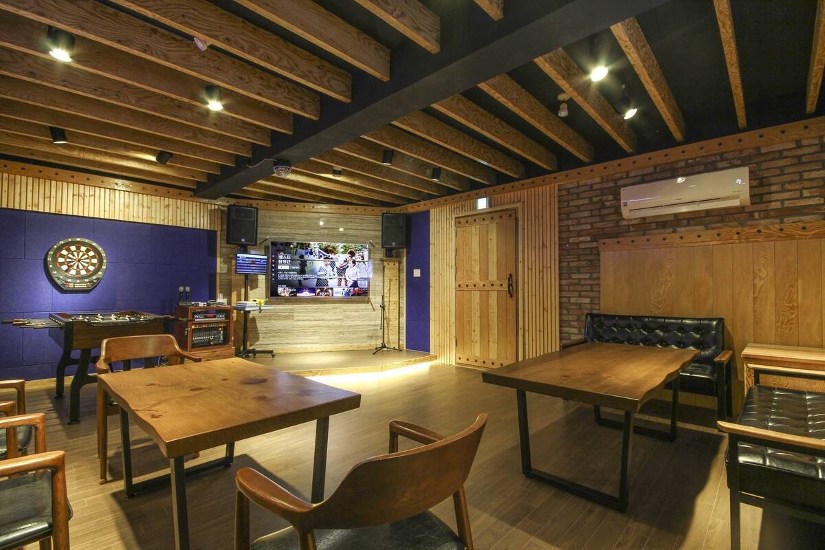 A restaurant interior with wooden tables and chairs, a dartboard, and a television screen displaying a sports event.