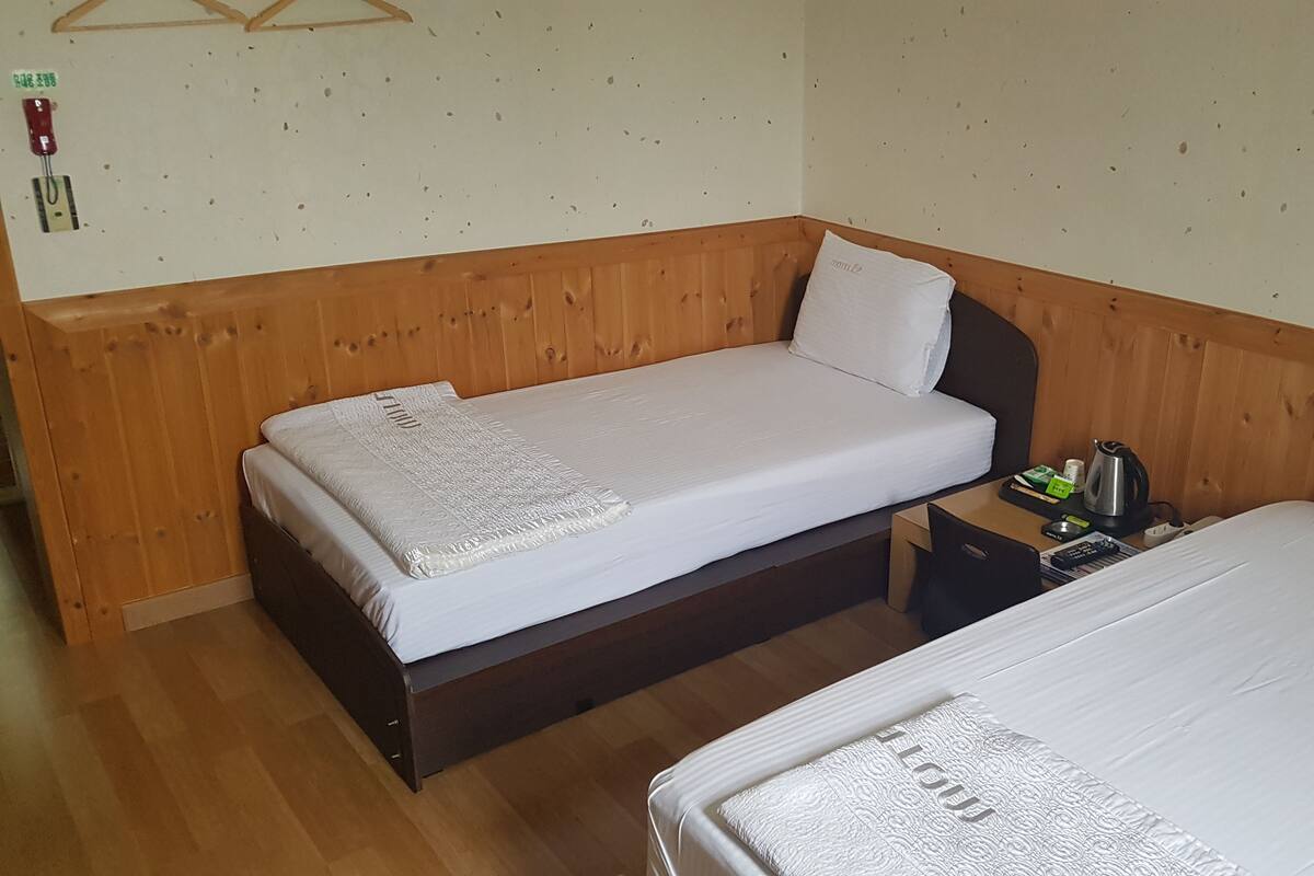 A small, cozy room with two single beds, wooden paneling on the walls, a small bedside table with a kettle and a remote control, and a wall-mounted fire alarm.