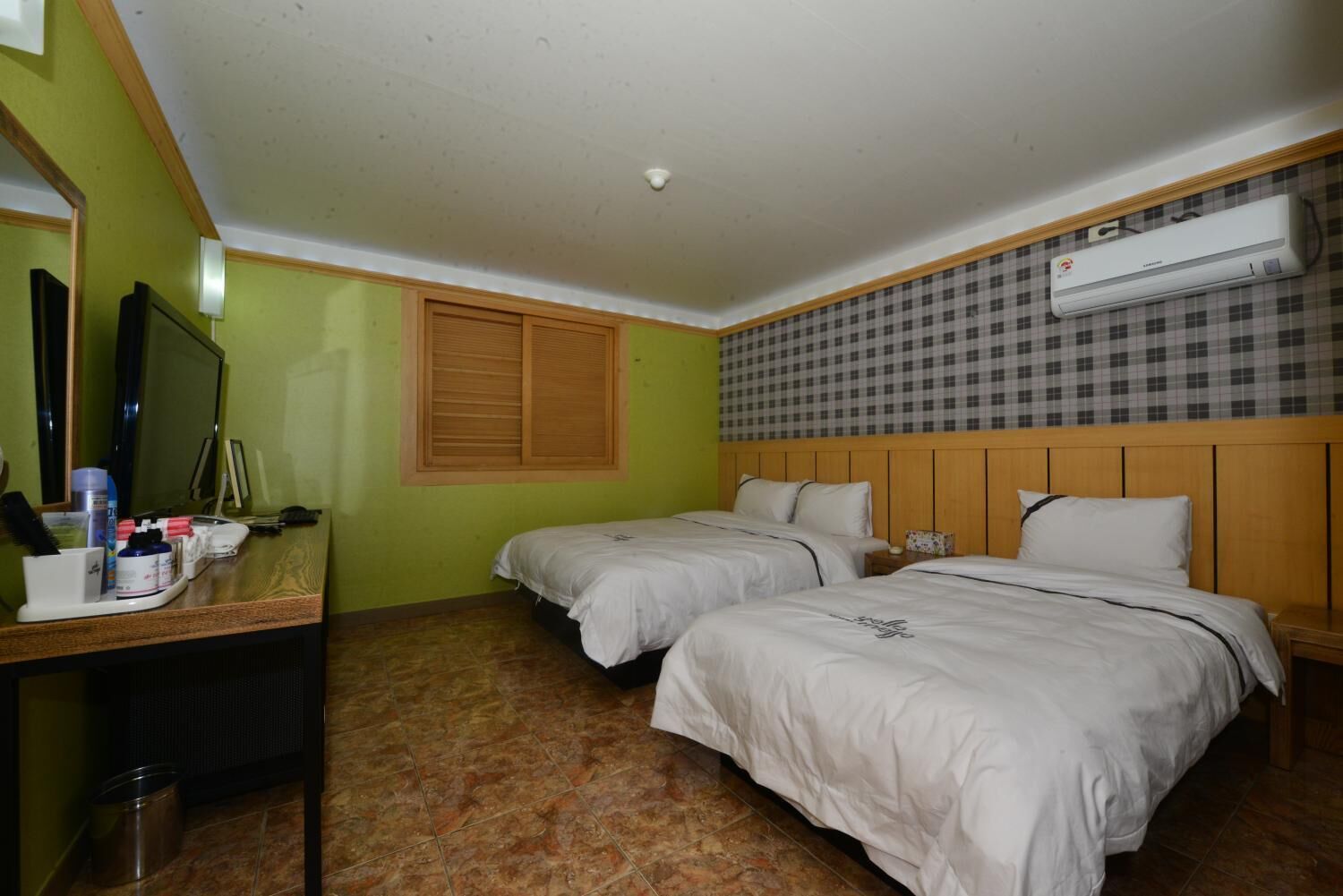 A hotel room with two beds, a desk with toiletries, a mirror, and an air conditioning unit.