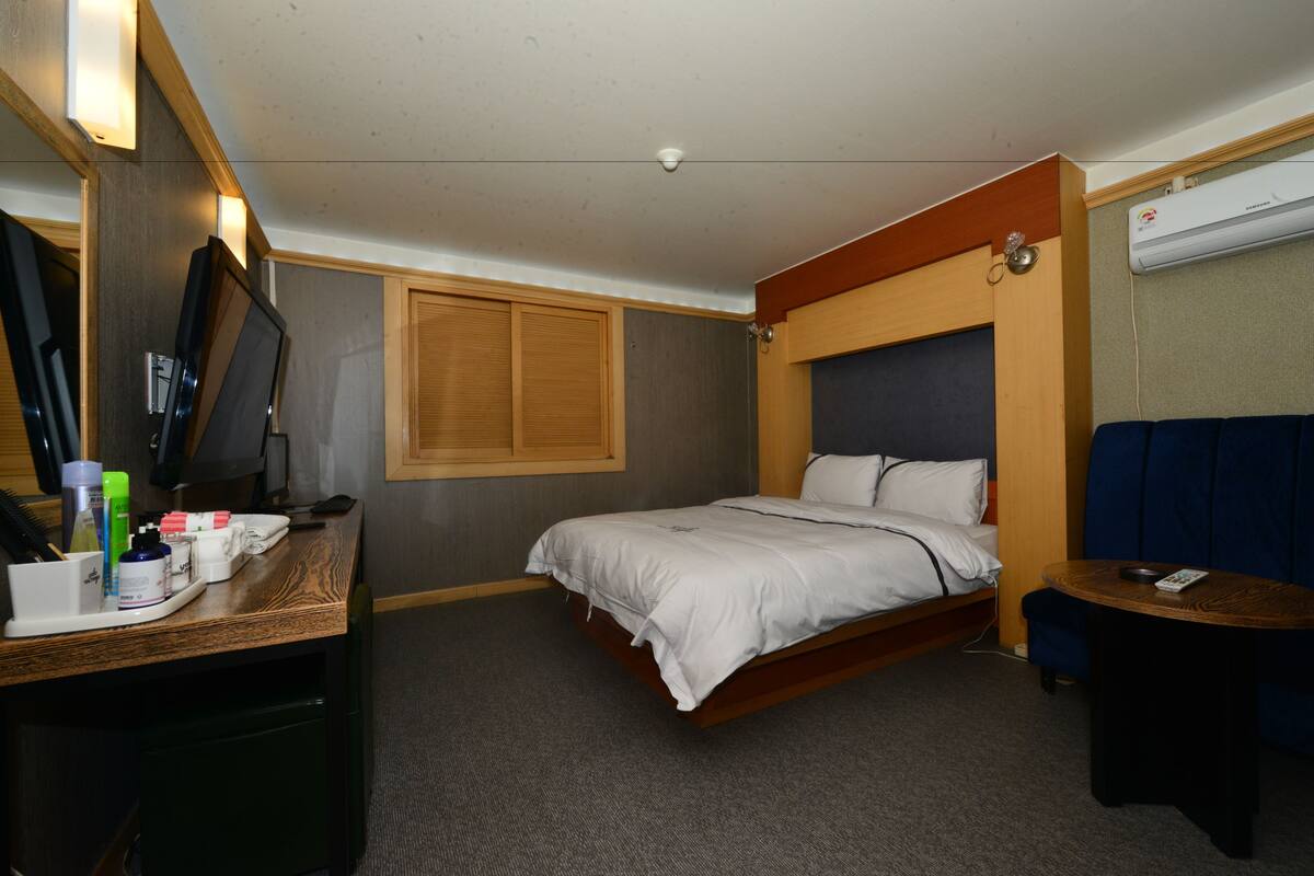 Ein kompaktes Hotelzimmer mit Bett, Schreibtisch mit Pflegeartikeln, einem kleinen Tisch und einer Wand-Klimaanlage.