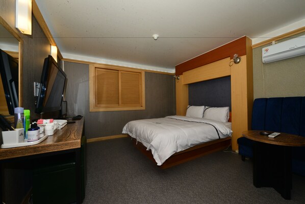 A compact hotel room with a bed, a desk with toiletries, a small table, and a wall-mounted air conditioner.