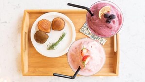 Una bandeja de madera con dos vasos de batidos rosados, uno con arándanos y otro con fresas, y un plato con tres galletas y una ramita de romero.