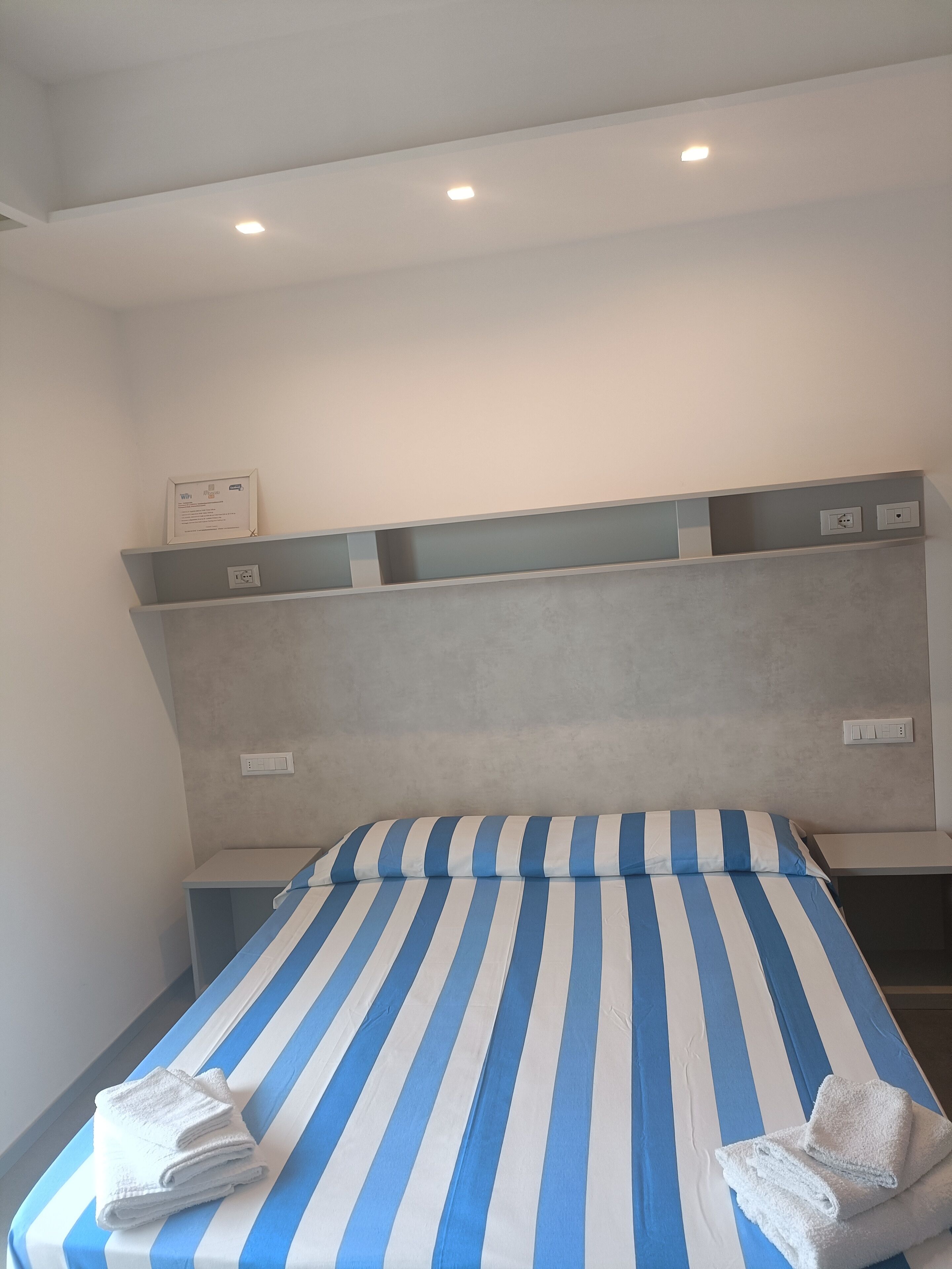 Una habitación de hotel con una cama y ropa de cama azul y blanca a rayas, un cabecero con estantes integrados y un techo con iluminación empotrada.