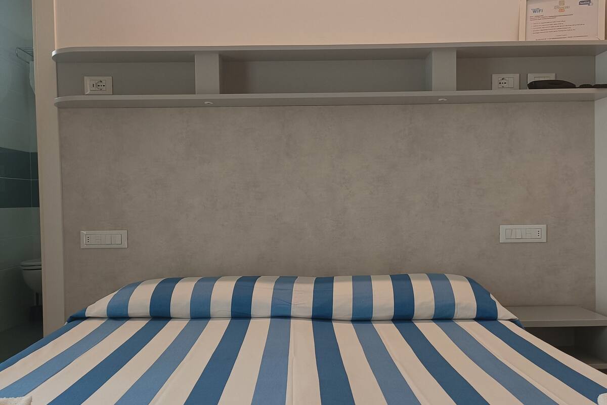 Una habitación de hotel con cama, cabecero con estantes integrados y lámpara fijada a la pared.