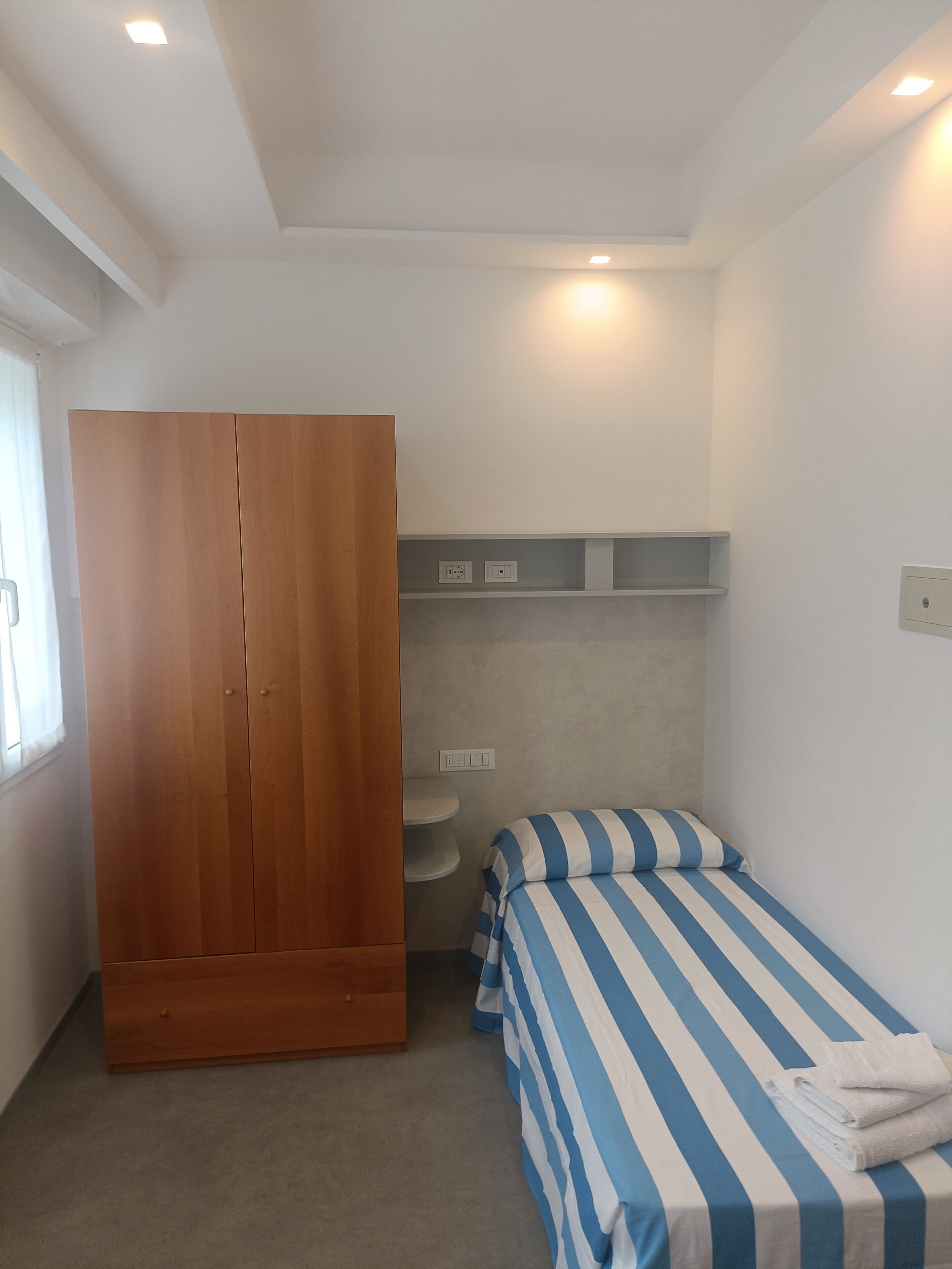 A bedroom with a wooden wardrobe, a bed with striped bedding, a bedside table, and a shelf unit.