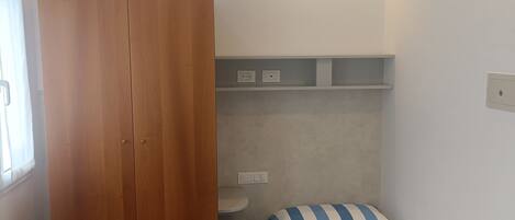 A bedroom with a wooden wardrobe, a bed with striped bedding, a bedside table, and a shelf unit.