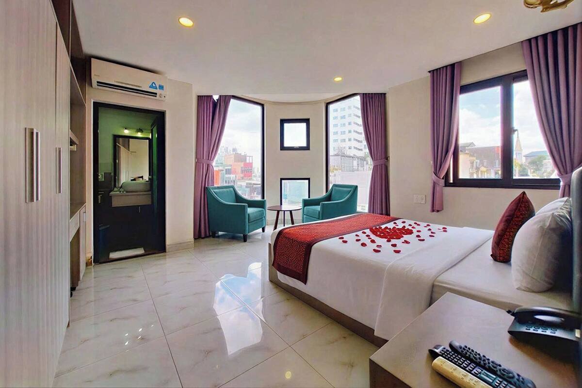 A modern hotel room with a large bed, two chairs, a small table, and a view of the city through large windows.
