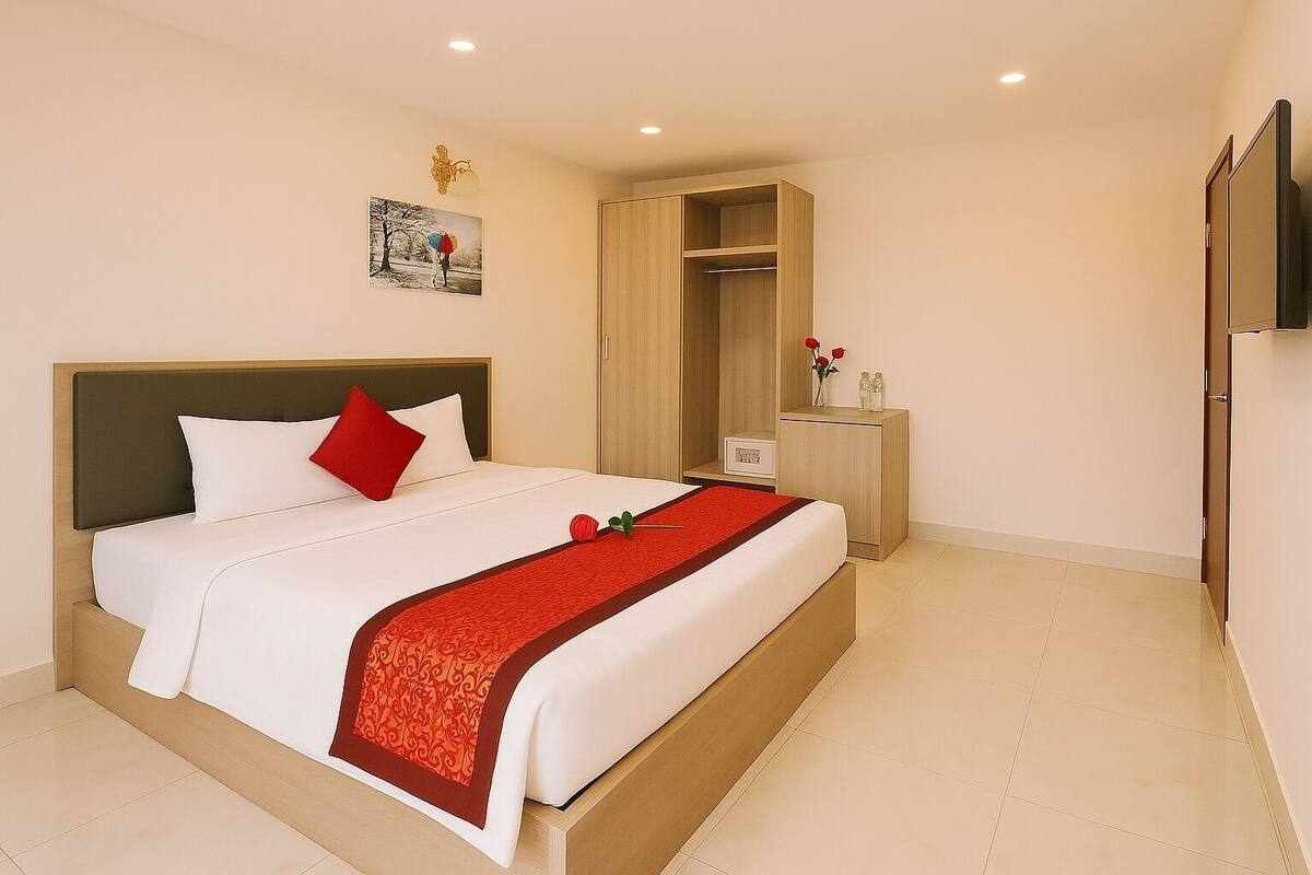 A neatly made bed with white bedding and a red accent, a bedside table with a vase of red roses, and a wooden wardrobe.