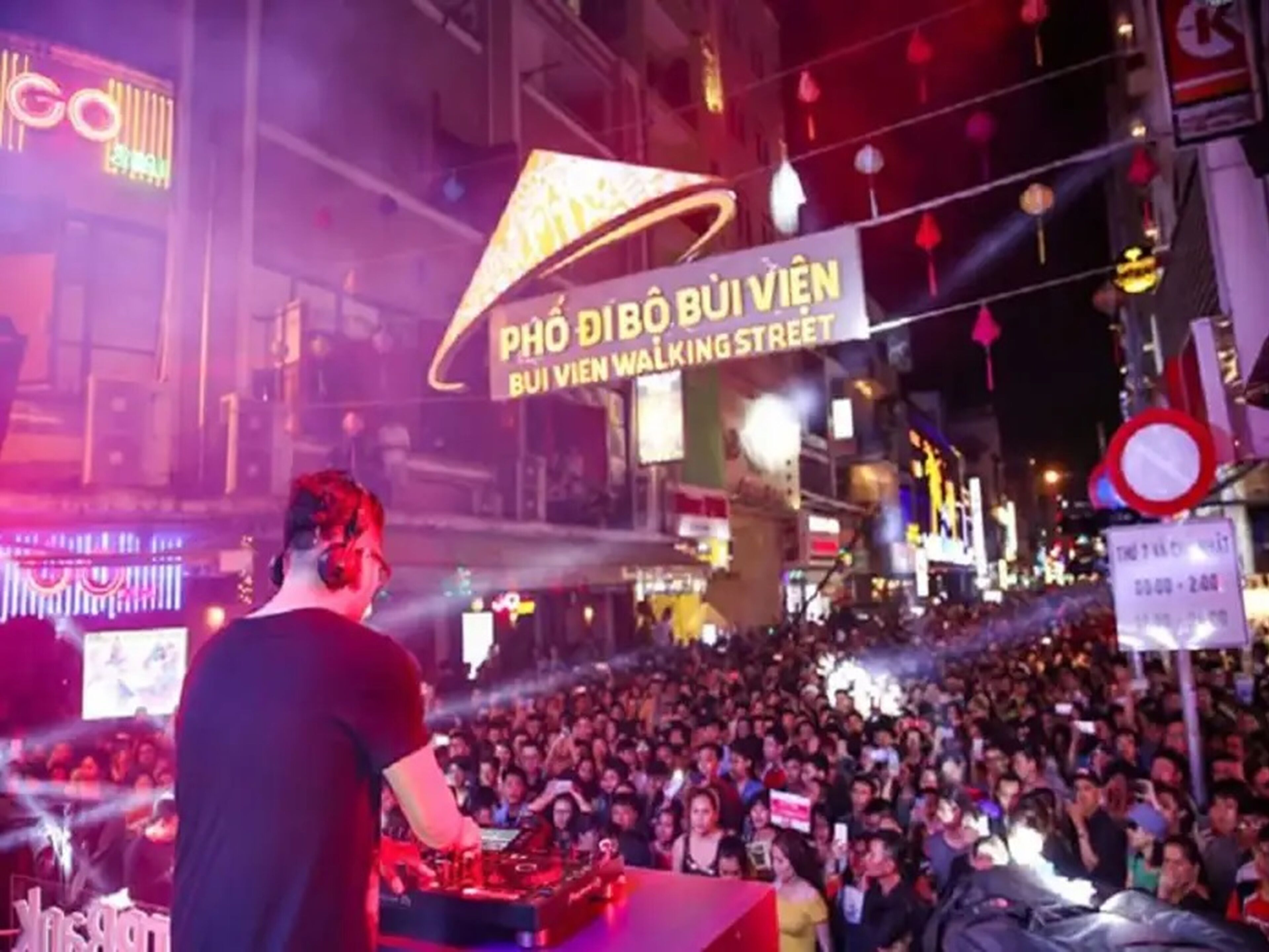 A crowded street scene with a DJ booth and a large illuminated sign.