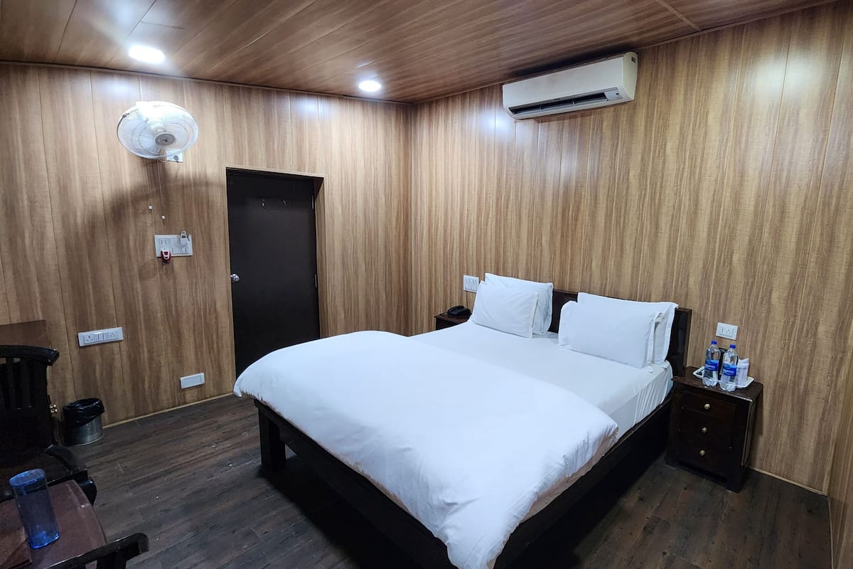 A hotel room with a wooden paneled wall, a bed with white bedding, a bedside table with a lamp, and an air conditioning unit.