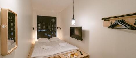 Un dormitorio pequeño y ordenado con cama, mesita de noche y espejo. Se ve una ventana con vista a edificios por la noche, un televisor de pared y una lámpara colgante.