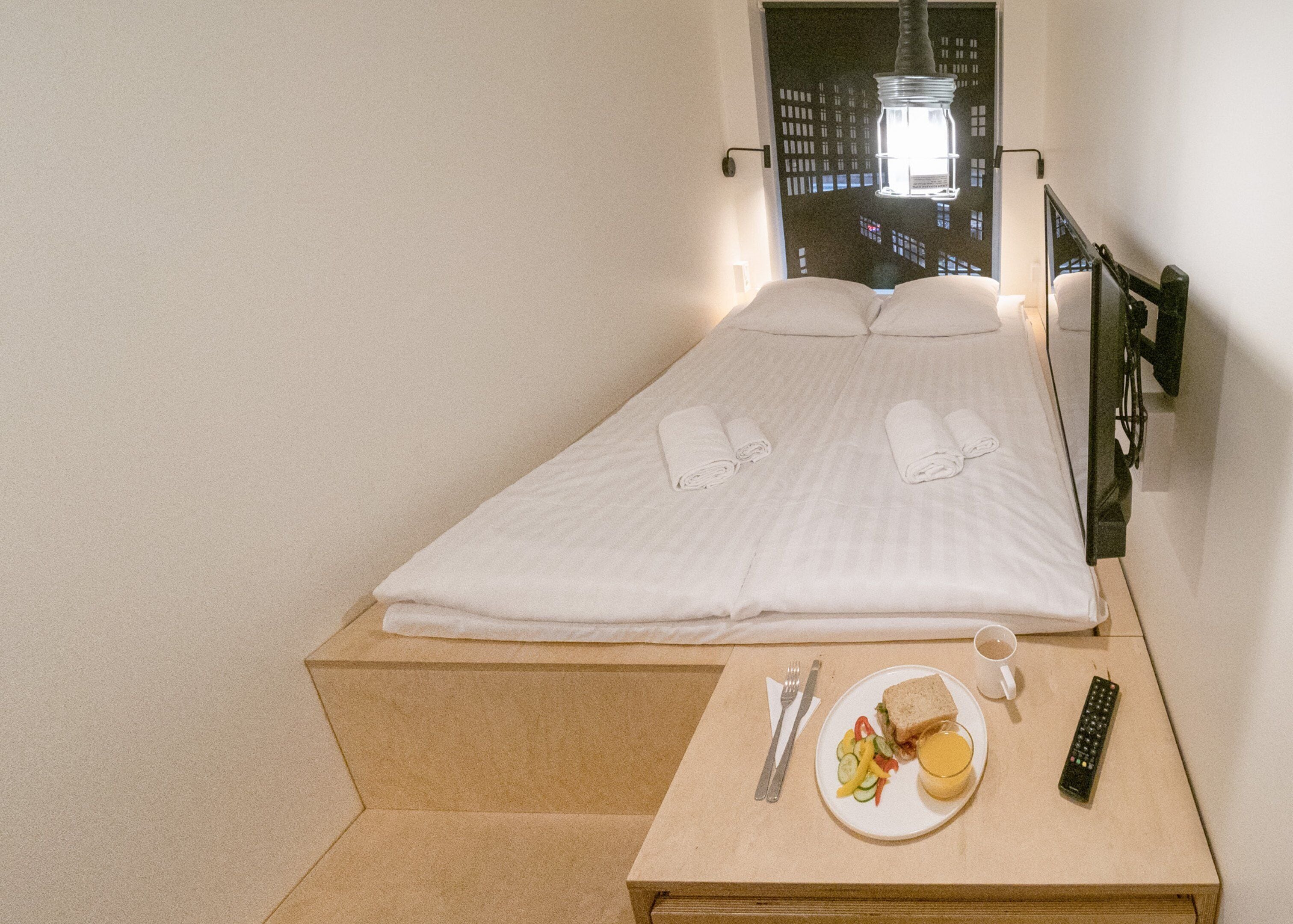 A neatly made bed with white linens and towels, a bedside table with a plate of food, a cup, cutlery, and a remote control.
