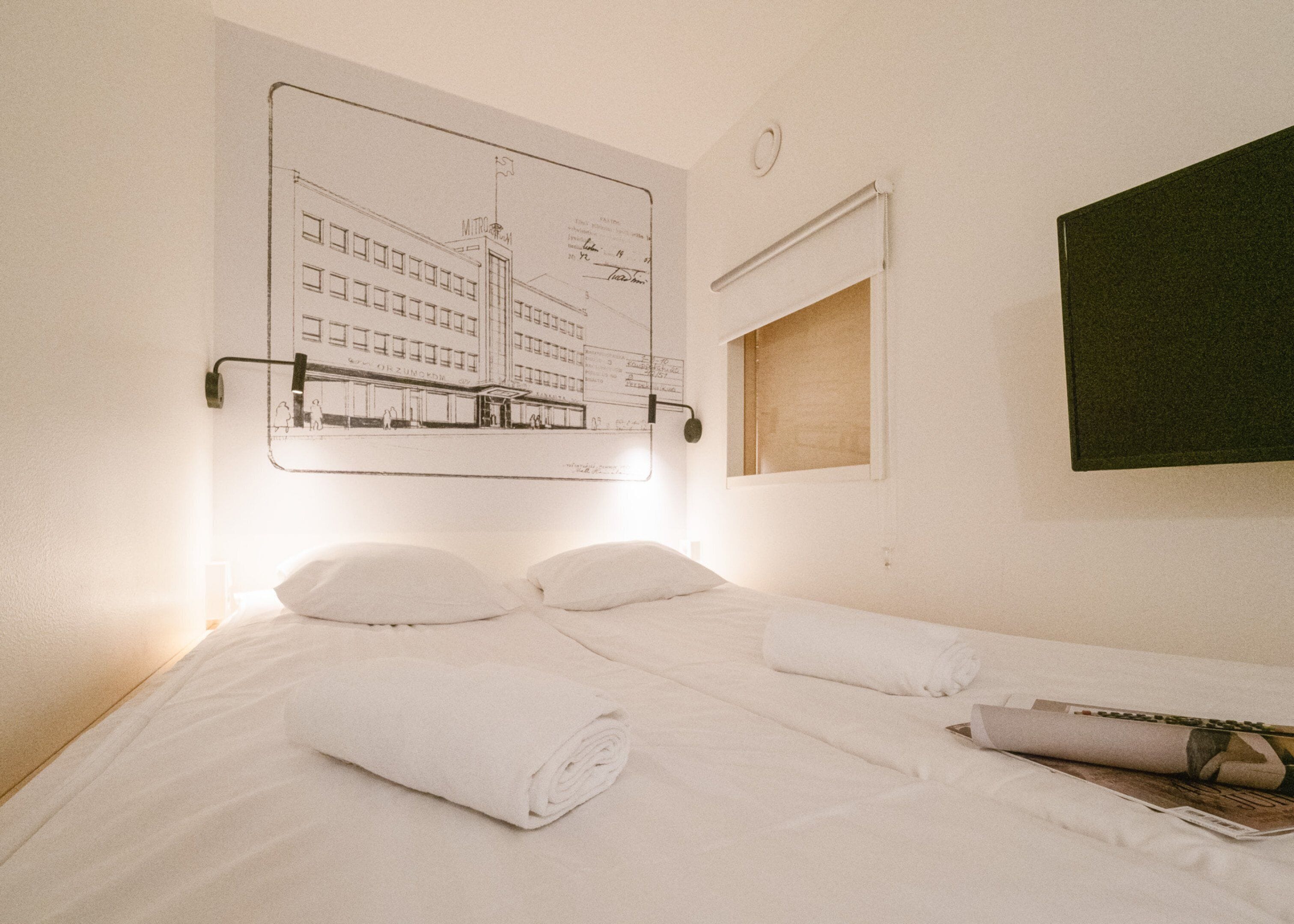 A hotel room with a bed, a wall-mounted light, a window, and a framed architectural drawing.