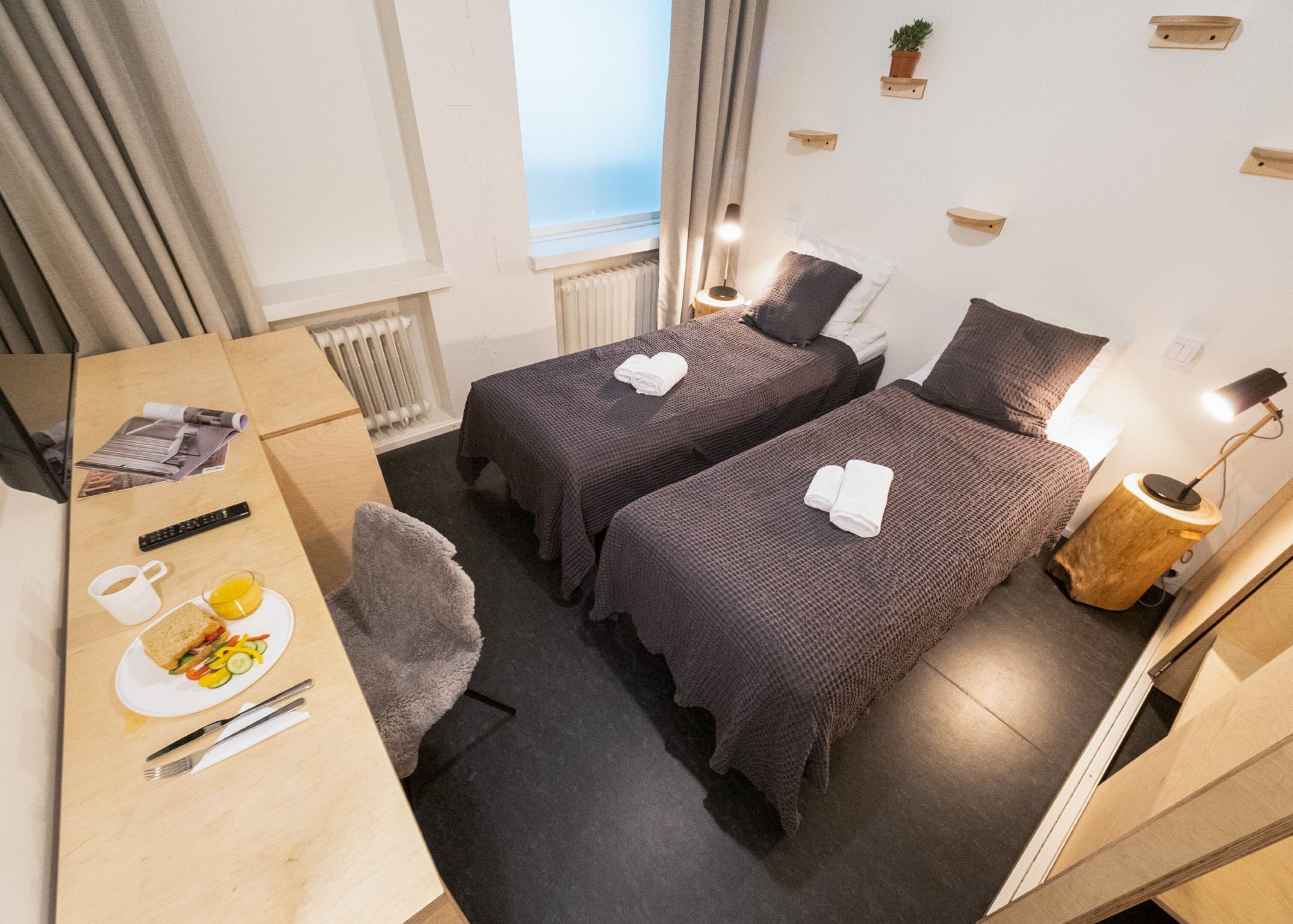 A hotel room with two beds, a desk with breakfast, a radiator, and a window with curtains.