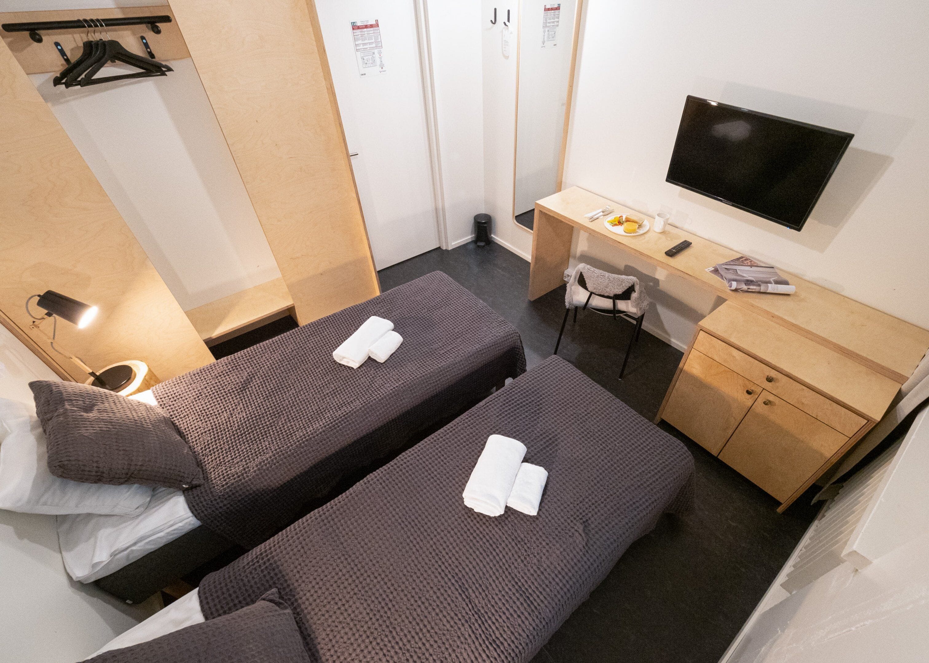 A compact hotel room with two single beds, a desk with a chair, a TV, and a small wooden cabinet.