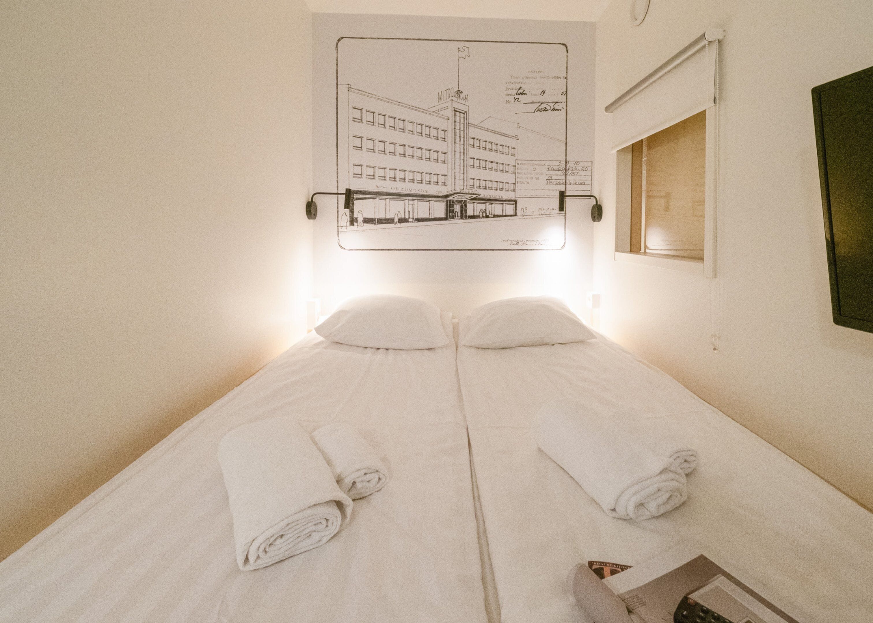 Un lit simple avec deux serviettes enroulées, une lampe fixée au mur et un plan d’architecture encadré.
