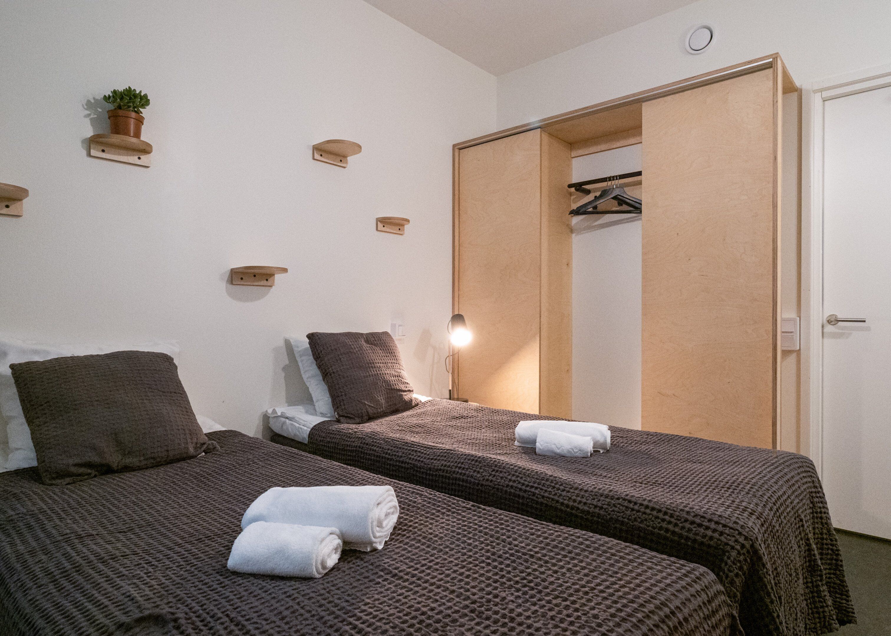 Habitació amb dos llits, cadascun amb llençol marró i almoades, una petita planta en testos a la paret, i un armari de fusta amb mirall.