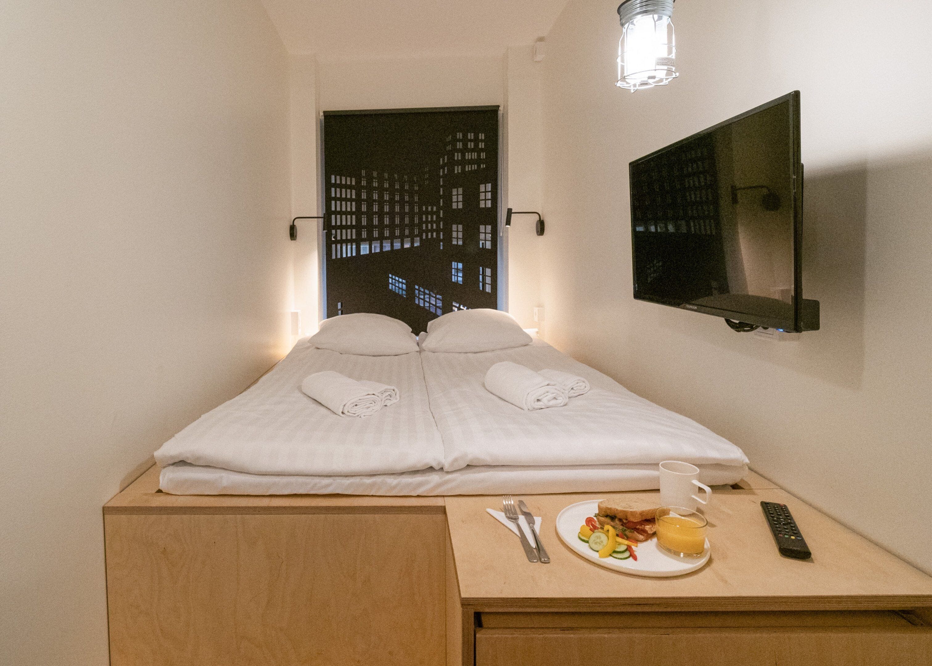 Quarto de hotel com uma cama, uma pequena escrivaninha com um prato de comida, um controle remoto e uma TV fixada na parede.
