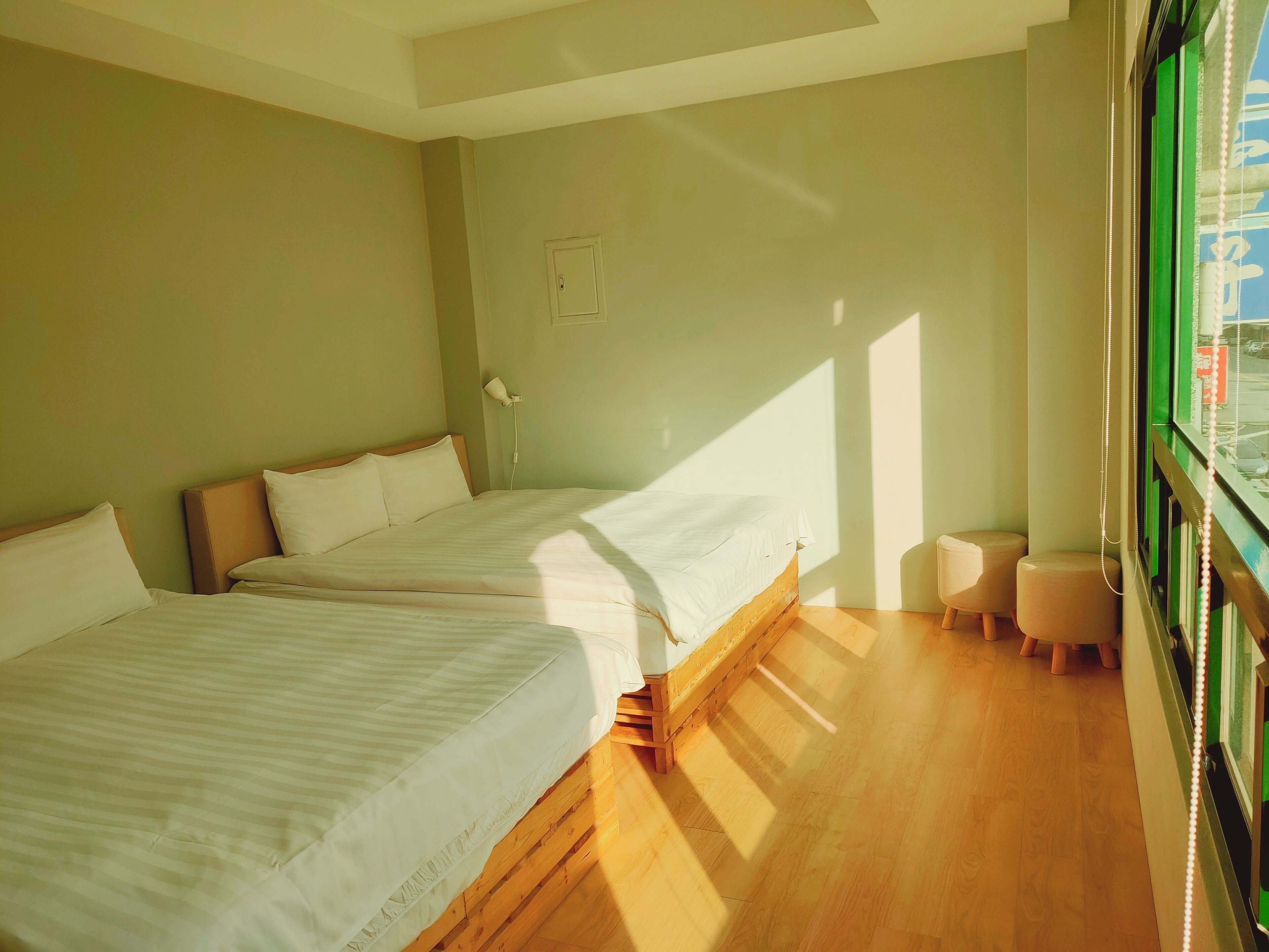 A room with two beds, a small stool, a wall socket, and a window with blinds.