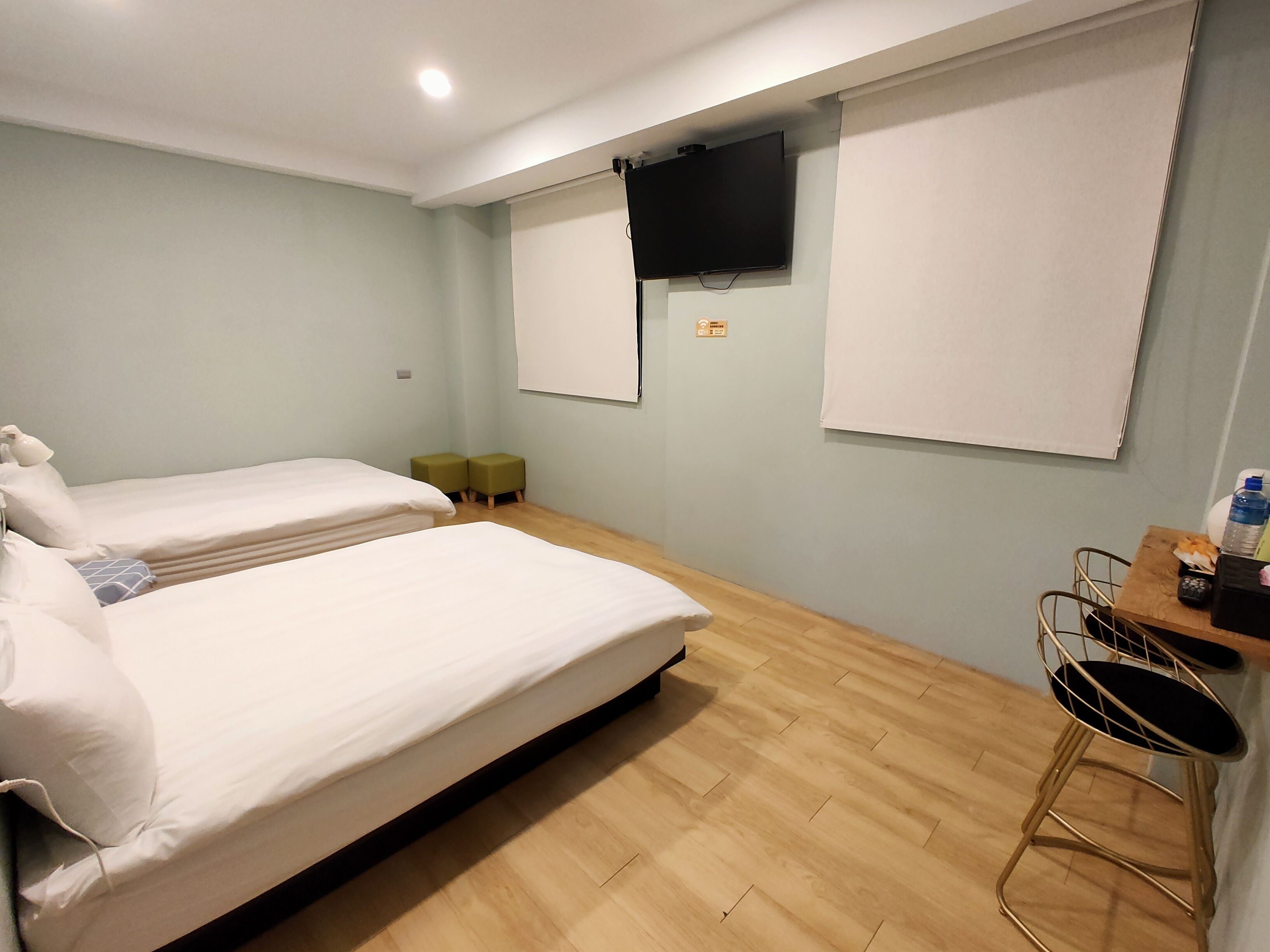 Habitación de hotel con dos camas, televisión, escritorio con silla y ventana con persianas.