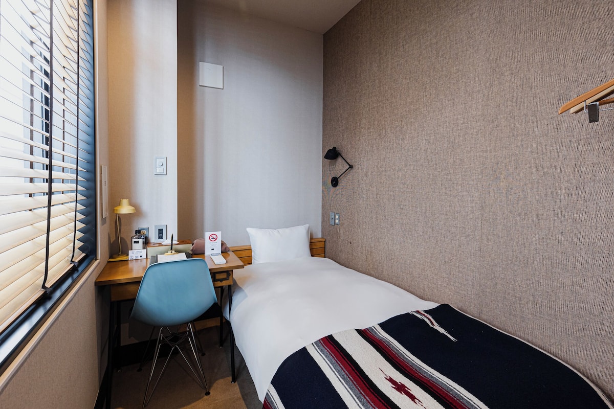 A hotel room with a bed, a desk with a chair, a lamp, and a window with blinds.