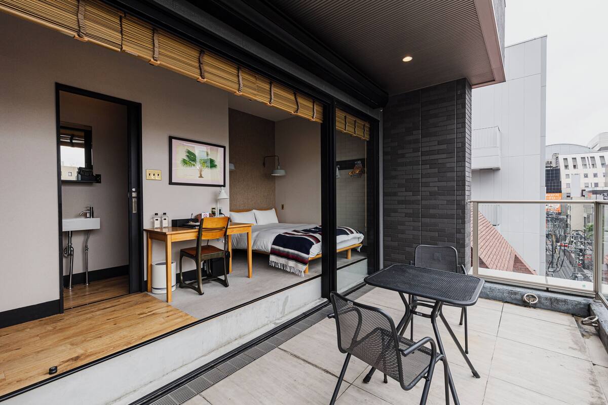 A modern room with a bed, desk, and chair, a small bathroom, and a balcony with a table and chairs.