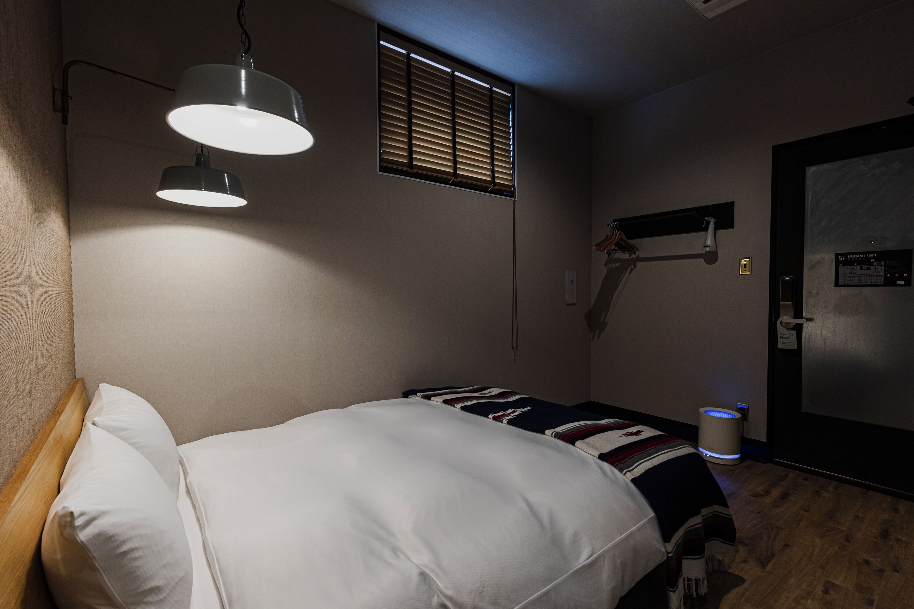 A bedroom with a bed, two pendant lights, a window with blinds, a door, and a wall-mounted shelf.