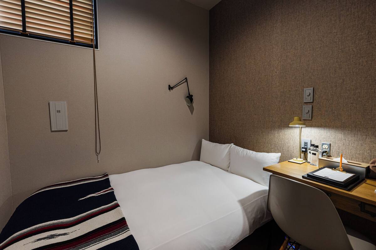 A hotel room with a single bed, a desk with a lamp, and a chair.