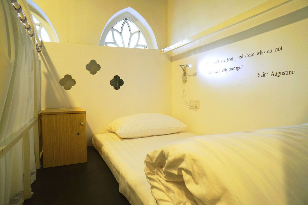 Une chambre à coucher comprenant un lit, une table de chevet, une fenêtre et une citation de Saint Augustin inscrite sur un mur.