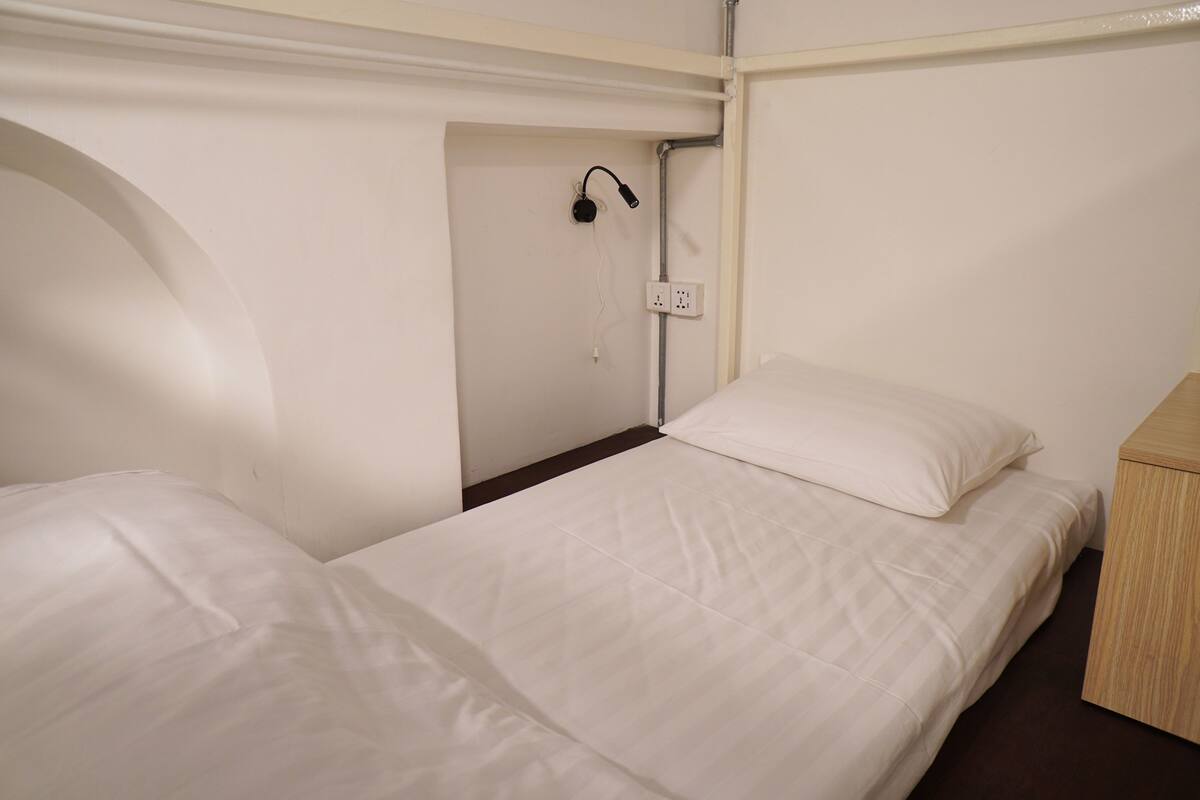 Une petite chambre propre, avec deux lits simples, une table de chevet en bois, une prise de courant et une lampe fixée au mur.