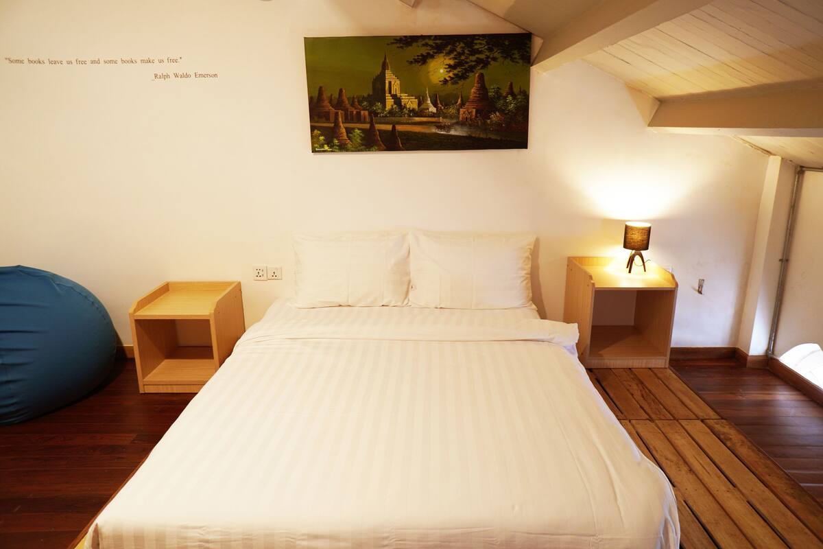 Un lit bien fait, avec du linge de lit blanc, deux tables de chevet avec des lampes, un fauteuil poire bleu et un tableau représentant un paysage accroché au mur.