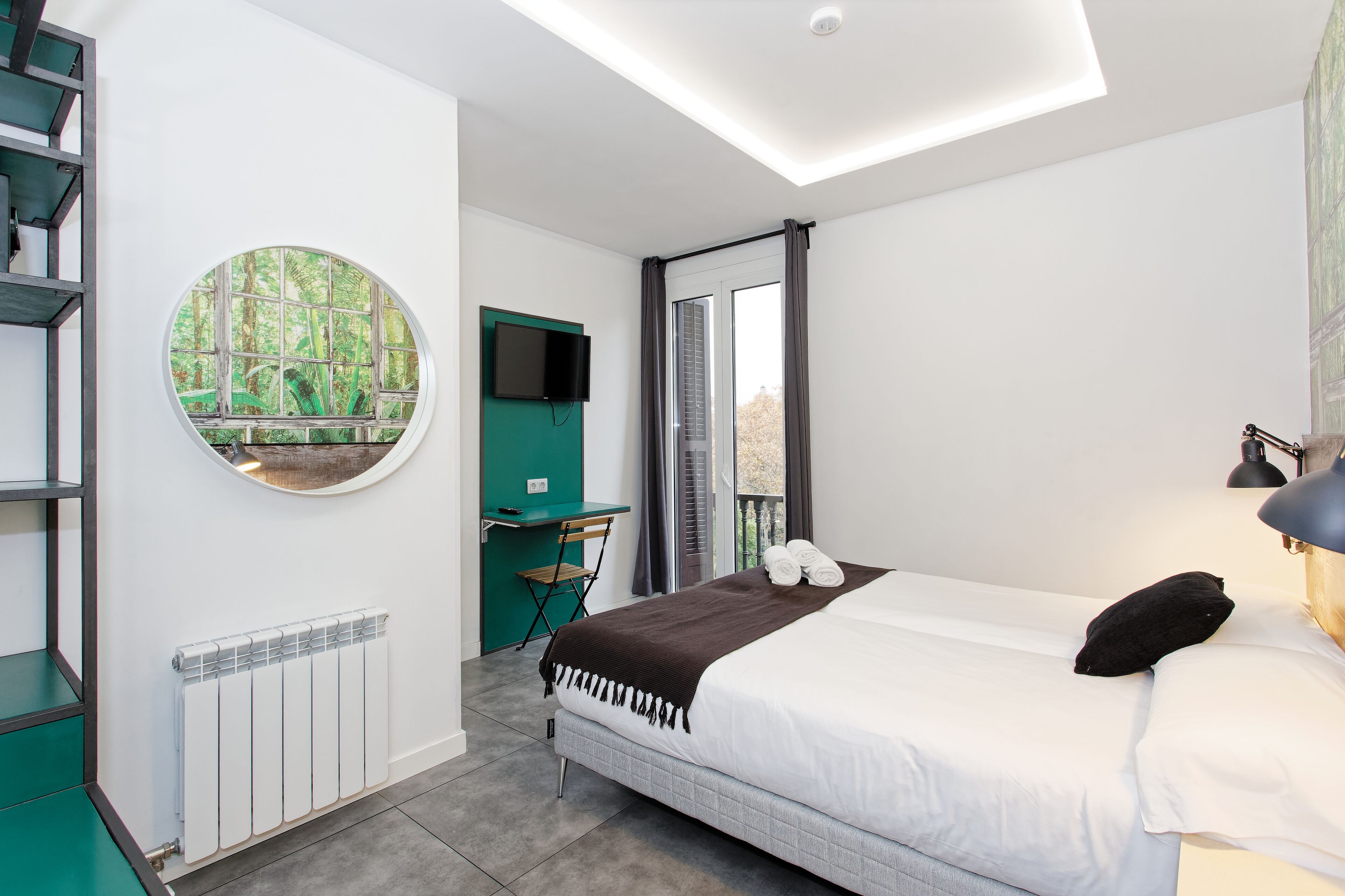 Quarto de hotel moderno com cama, escrivaninha, cadeira, espelho redondo e vista para o verde através de uma janela.