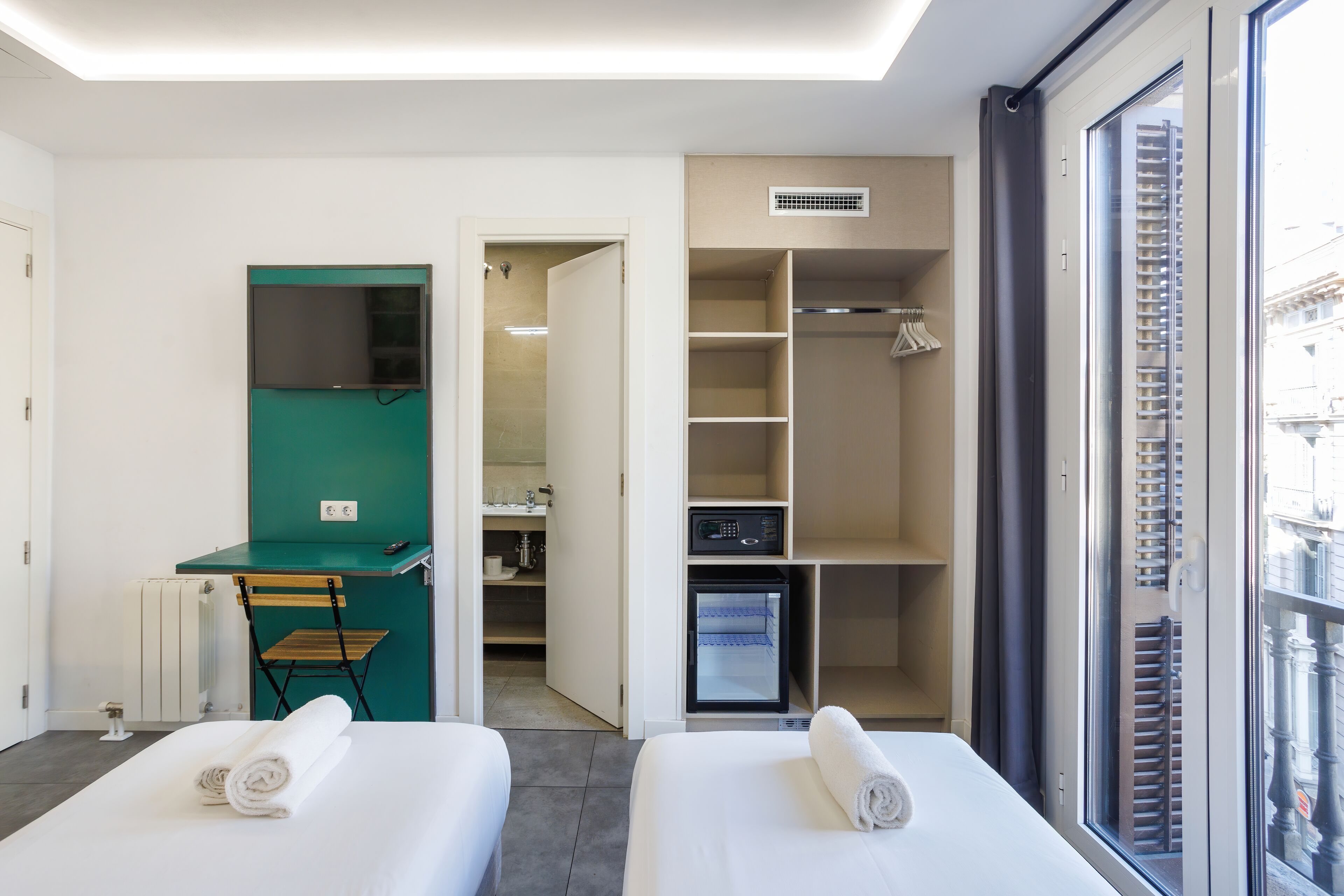 Habitación de hotel con dos camas, un escritorio, una silla, un minibar y vistas a la ciudad a través de un amplio ventanal.