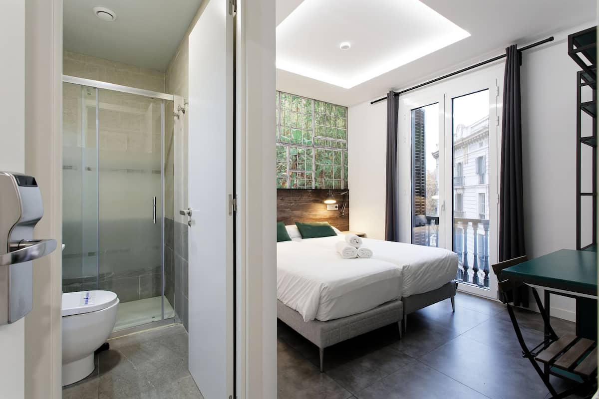 A modern hotel room with a glass-enclosed shower, a bed with white linens, a small table, and a view of a building through a large window.