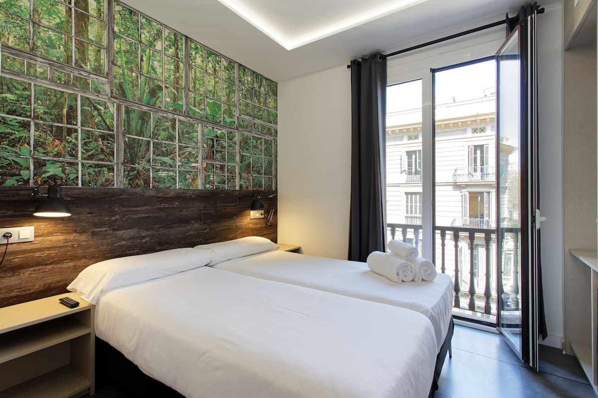 A hotel room with two beds, a wooden headboard, a large window with a view of buildings, and a balcony.