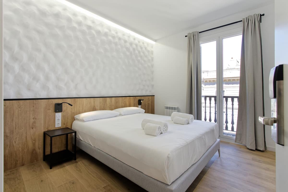 A modern hotel room with a large bed, a bedside table, a wall-mounted heater, and a view of the outside balcony.