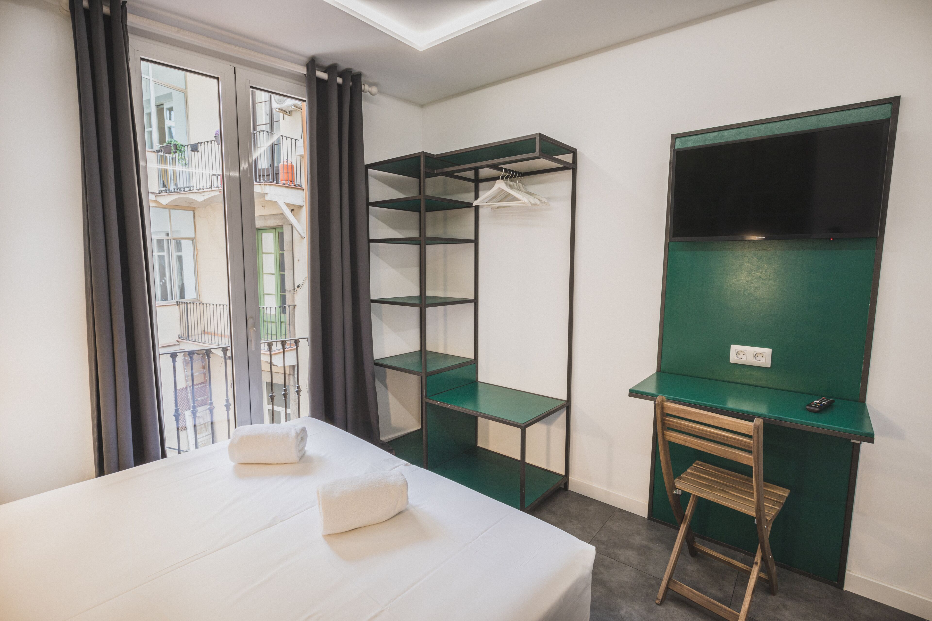 A hotel room with a bed, a green desk with a chair, a wardrobe, and a window with a view of a balcony.
