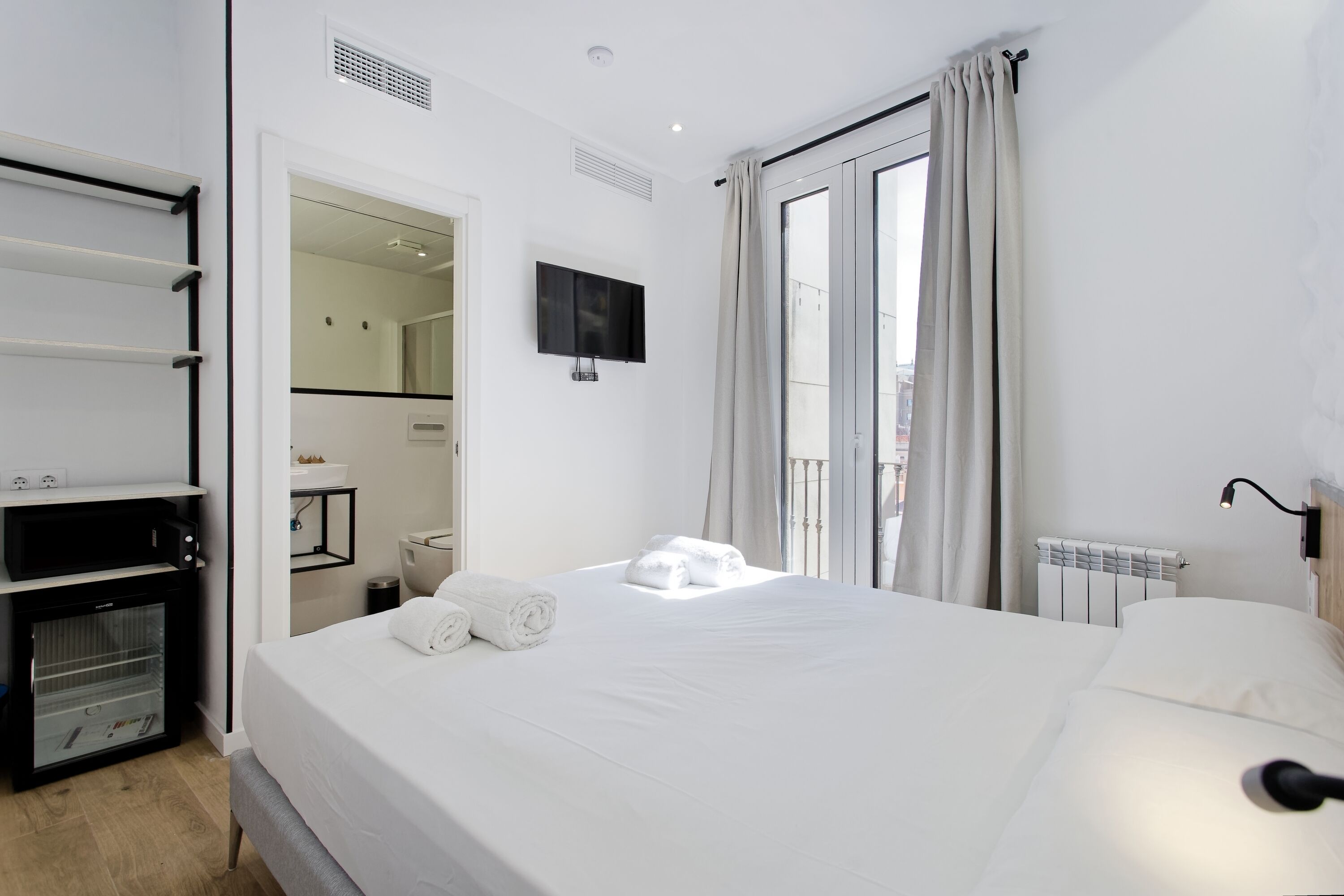 A hotel room with a bed, a TV, a minibar, and a view of the city through the window.