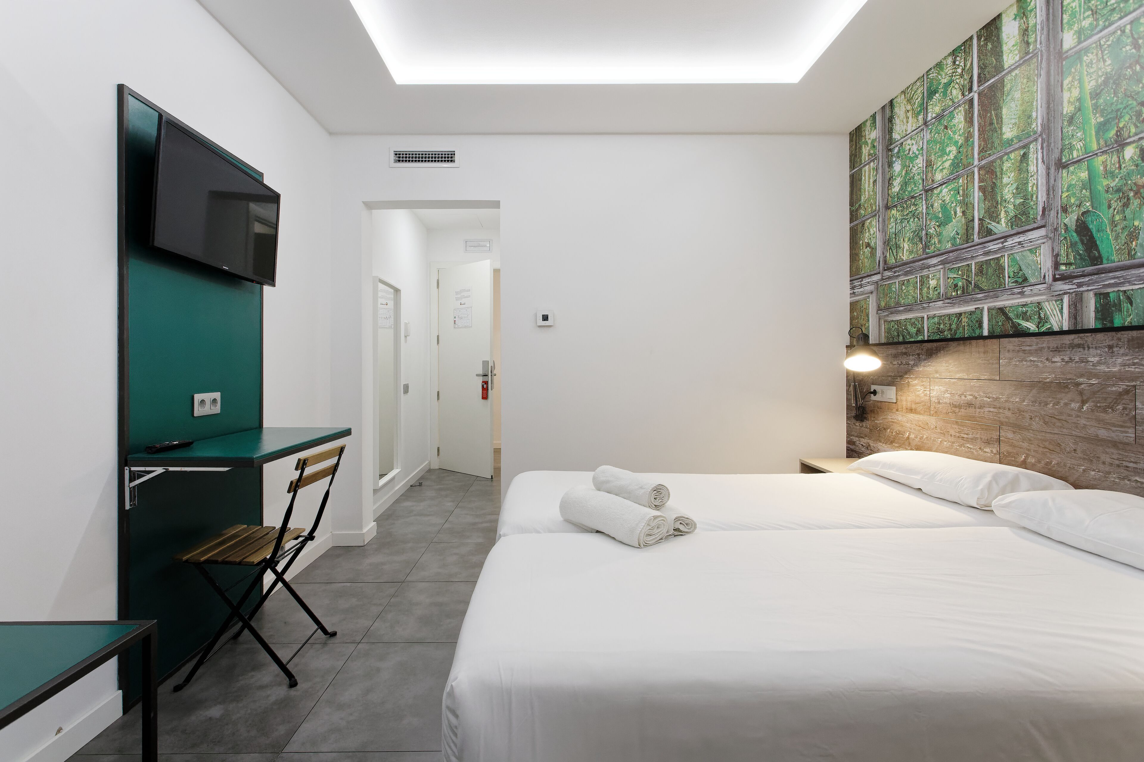 A hotel room with a bed, a desk, a chair, a flat-screen TV mounted on the wall, and a large window with a forest view.