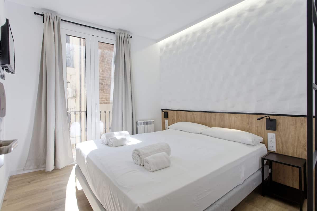 A modern hotel room with a large bed, a wooden headboard, a small bedside table, and a view of the city through the window.