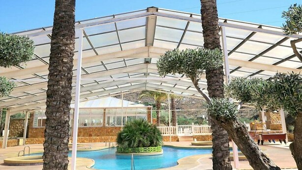 Un espace piscine couvert agrémenté de palmiers et doté d’une structure de toit en verre.