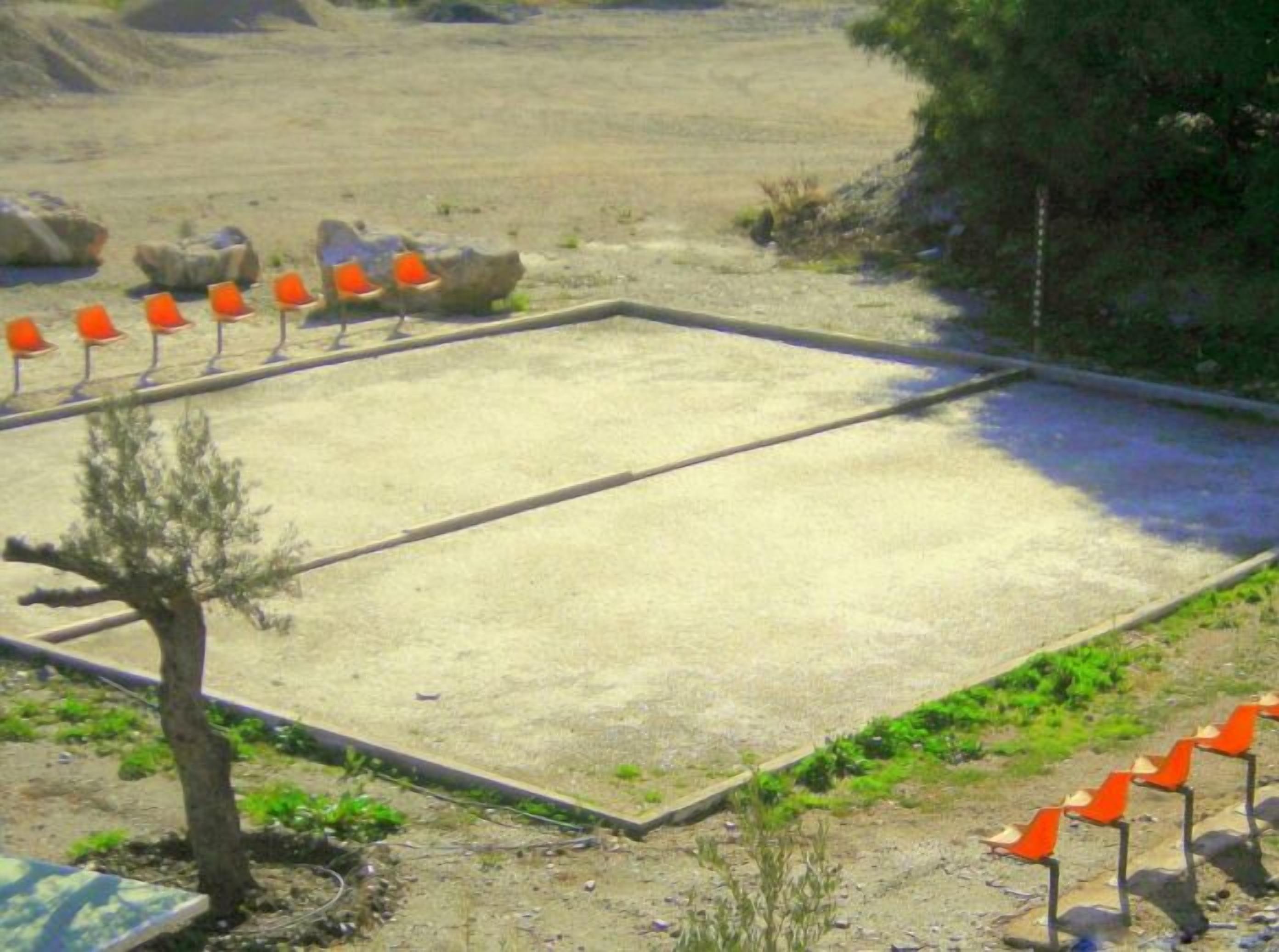 Un espace extérieur rectangulaire, doté d’une surface sablonneuse, entouré d’un petit muret en pierre et bordé d’une rangée de drapeaux orange.