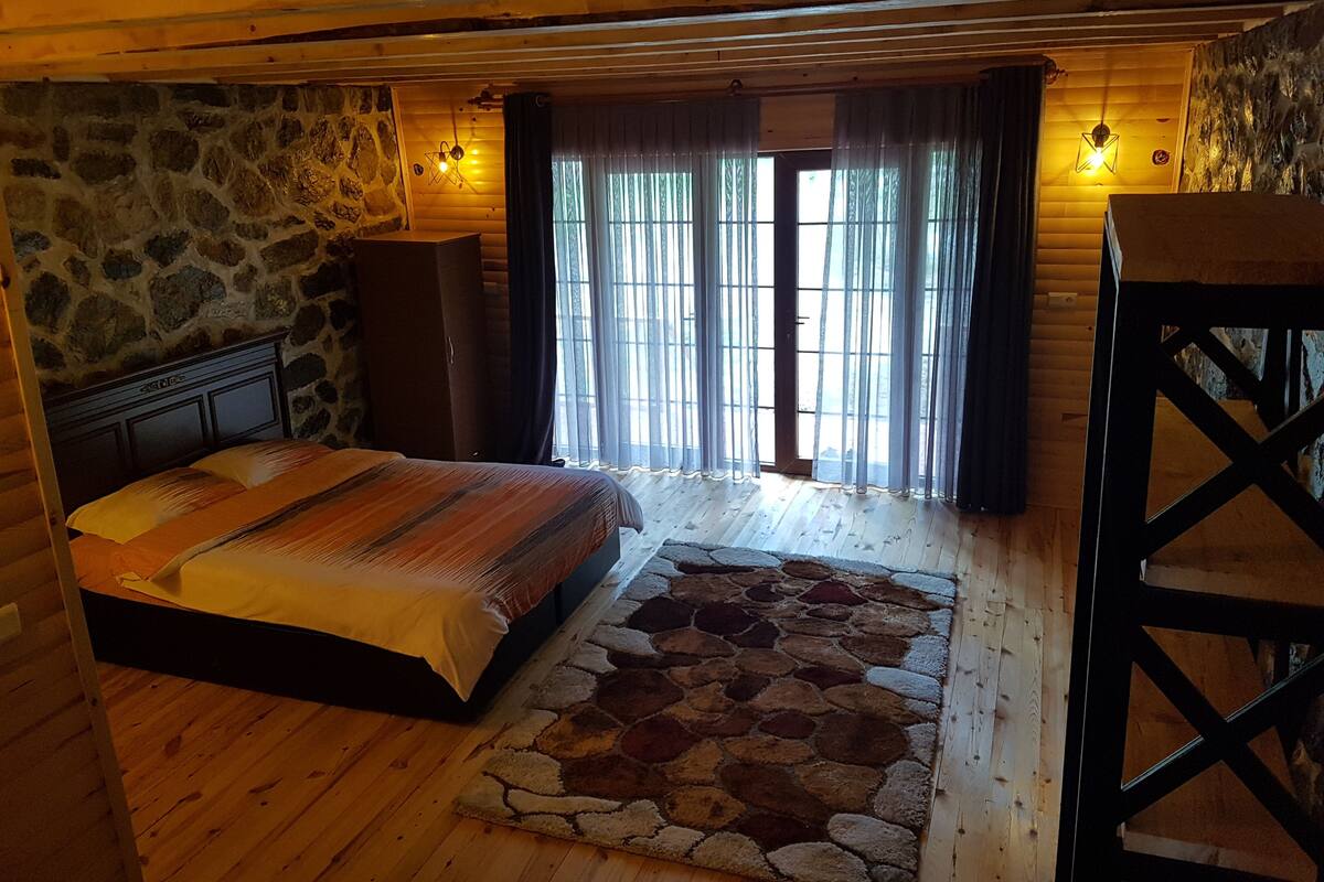 A wooden cabin bedroom with a stone wall, a bed with a striped blanket, a stone-patterned rug, and a wooden wardrobe.
