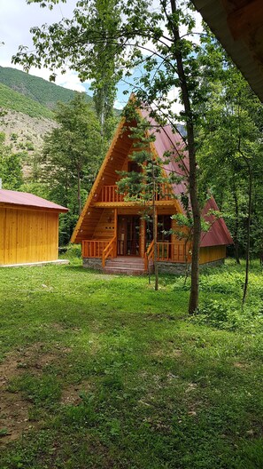 Una cabaña de madera con un porche y techo rojo, rodeada de exuberante vegetación y una colina al fondo.