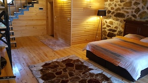 A wooden cabin interior with a staircase, a bed, a stone wall, and a stone-patterned rug.