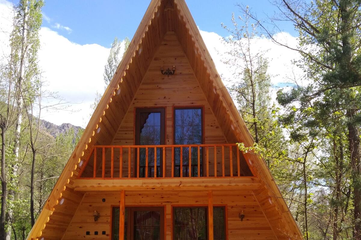 A wooden cabin with a balcony, stone foundation, and a view of the surrounding forest.