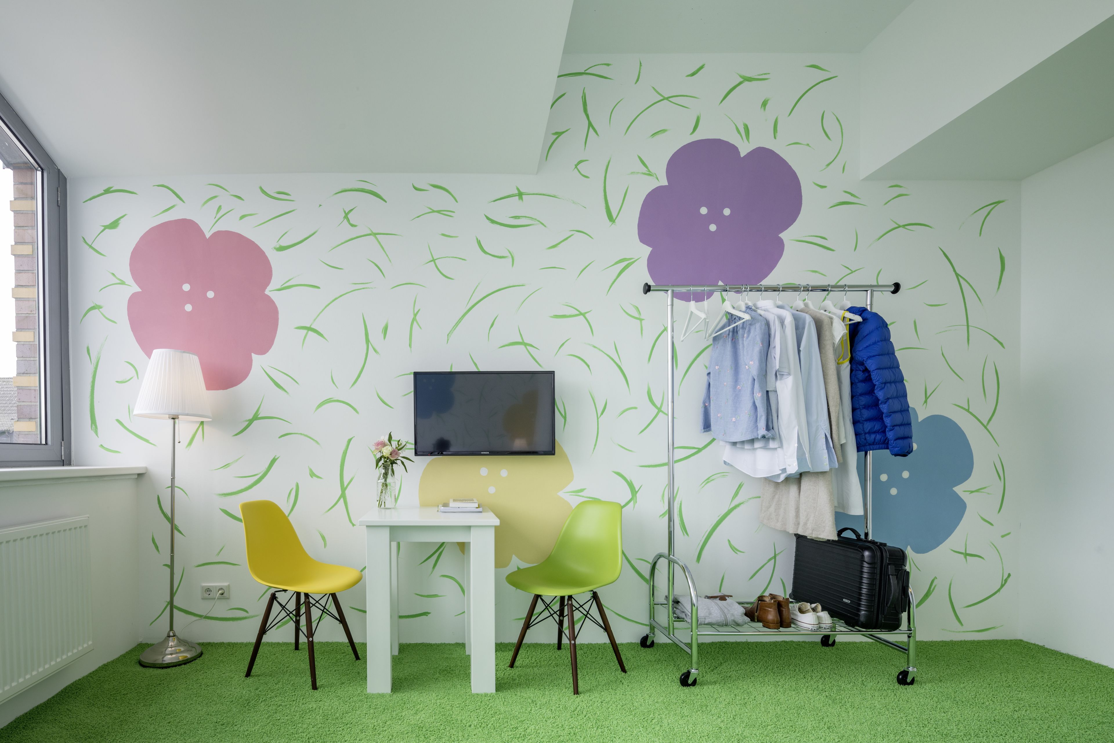 Una habitación con alfombra verde, una mesa blanca, dos sillas, un televisor y un perchero con ropa y una maleta.