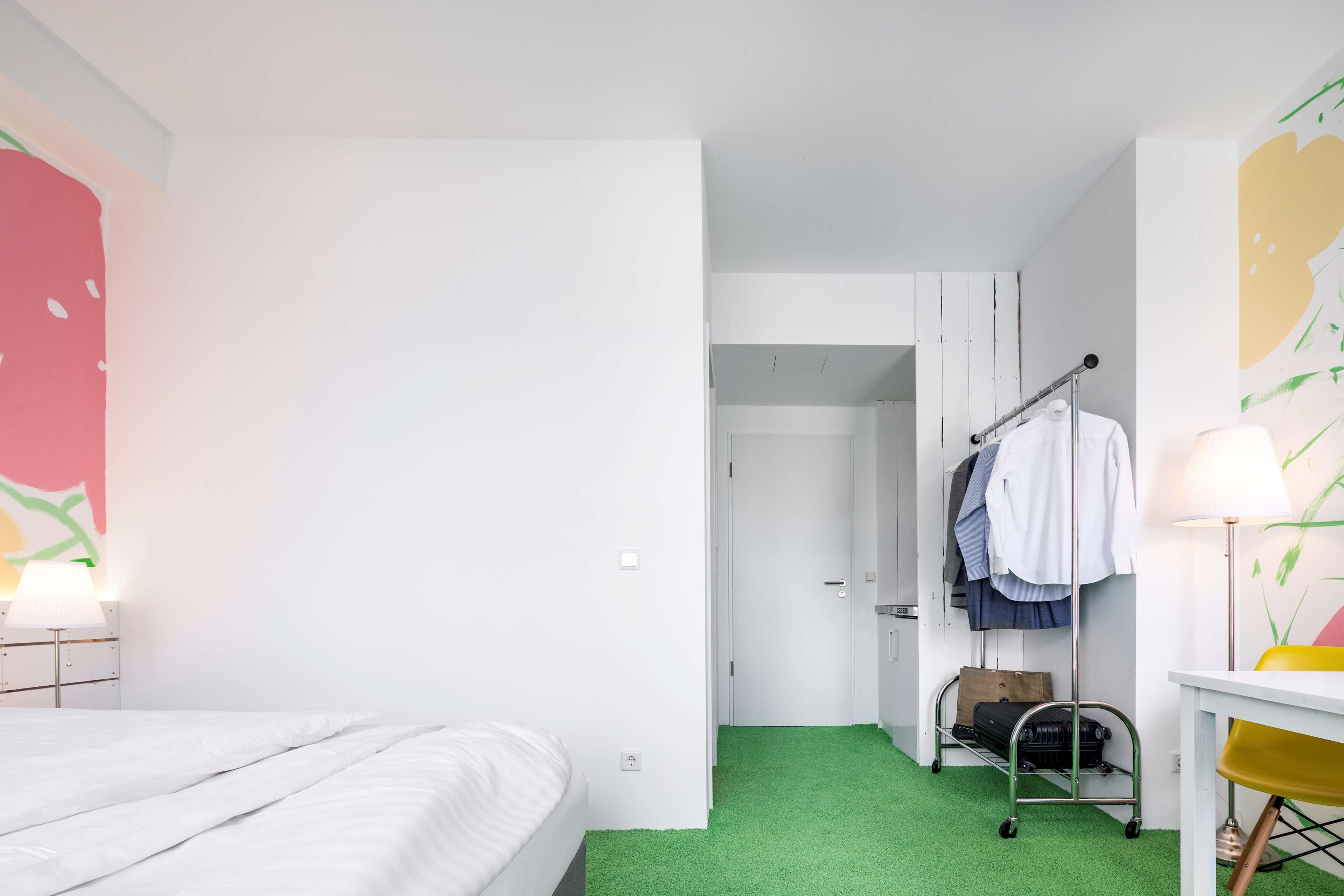 Une chambre d’hôtel avec un lit, un bureau, une chaise, une armoire et une valise posée sur un tapis vert.