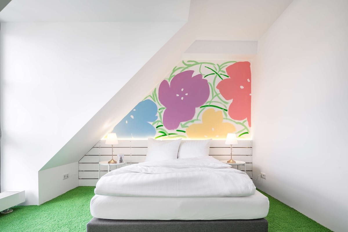 Ein Schlafzimmer mit schrägem Dach, einem Bett mit weißer Bettwäsche, einem farbenfrohen floralen Wandbild und einem Boden aus grünem Kunstgrün.
